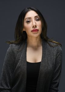 Nav Dhillon - founder of 1iCollective is wearing a black jacket with a black low neck rounded black t-shirt underneath. Her long black hair is loose around her and she is looking forward without her prosthetic eye in place on her left. Dark background.